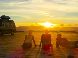 Drei junge Männer sitzen am Strand, die Sonne geht hinter Ihnen unter. Die Sonne taucht das Licht in einen goldenen Ton. Am linken Bildrand kann man einen Campervan erkennne, mit Dachbox. 