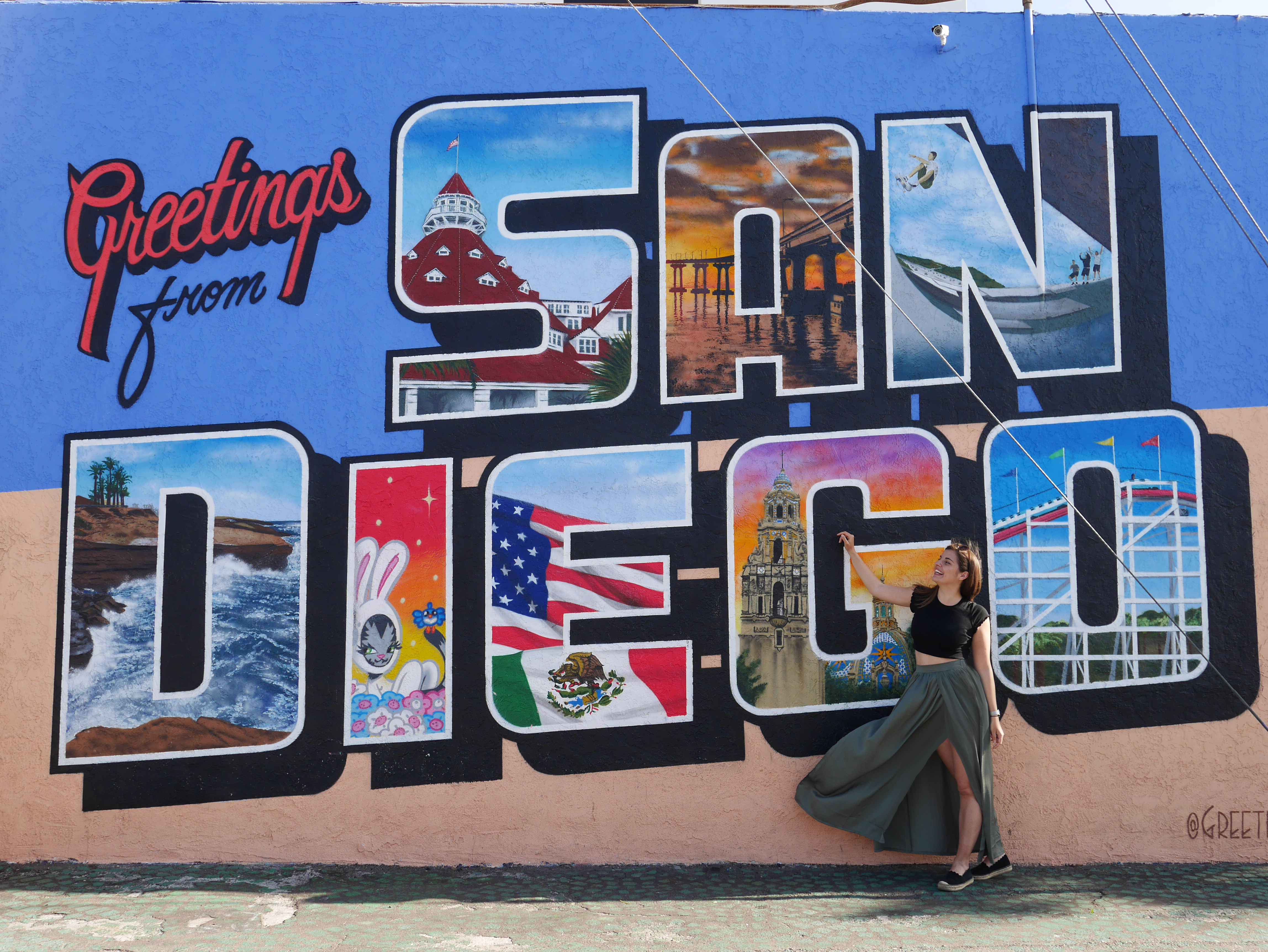 Eine junge Frau steht vor einer bunt bemalten Wand auf der "Greetings from San Diego" zu lesen ist. Sie trägt ein T-Shirt und einen langen Rock, der im Wind weht.